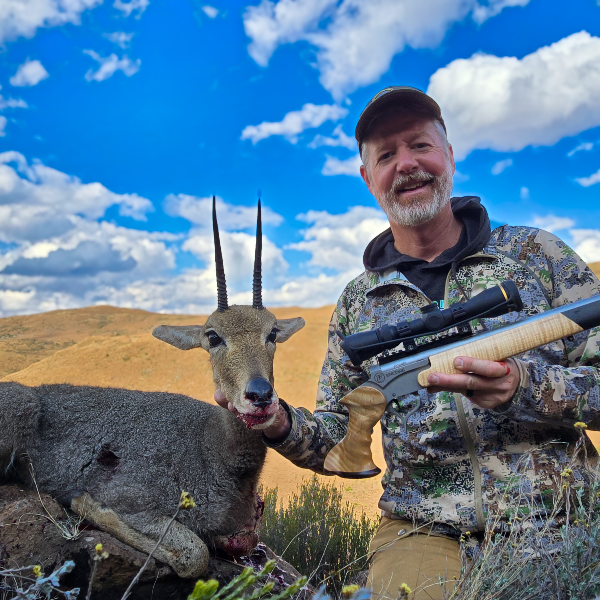 Hunter holding a grey rhebok ram and a scoped Encore handgun on a high grass slope