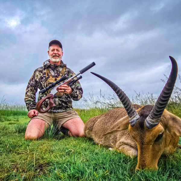 Hunter kneeling in green grass with a reedbuck and a suppressed handgun