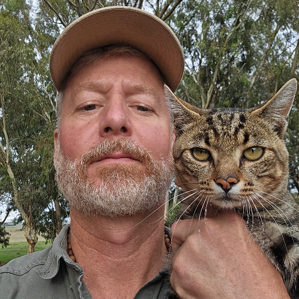 Tabby cat held up beside a bearded man in a cap under gum trees