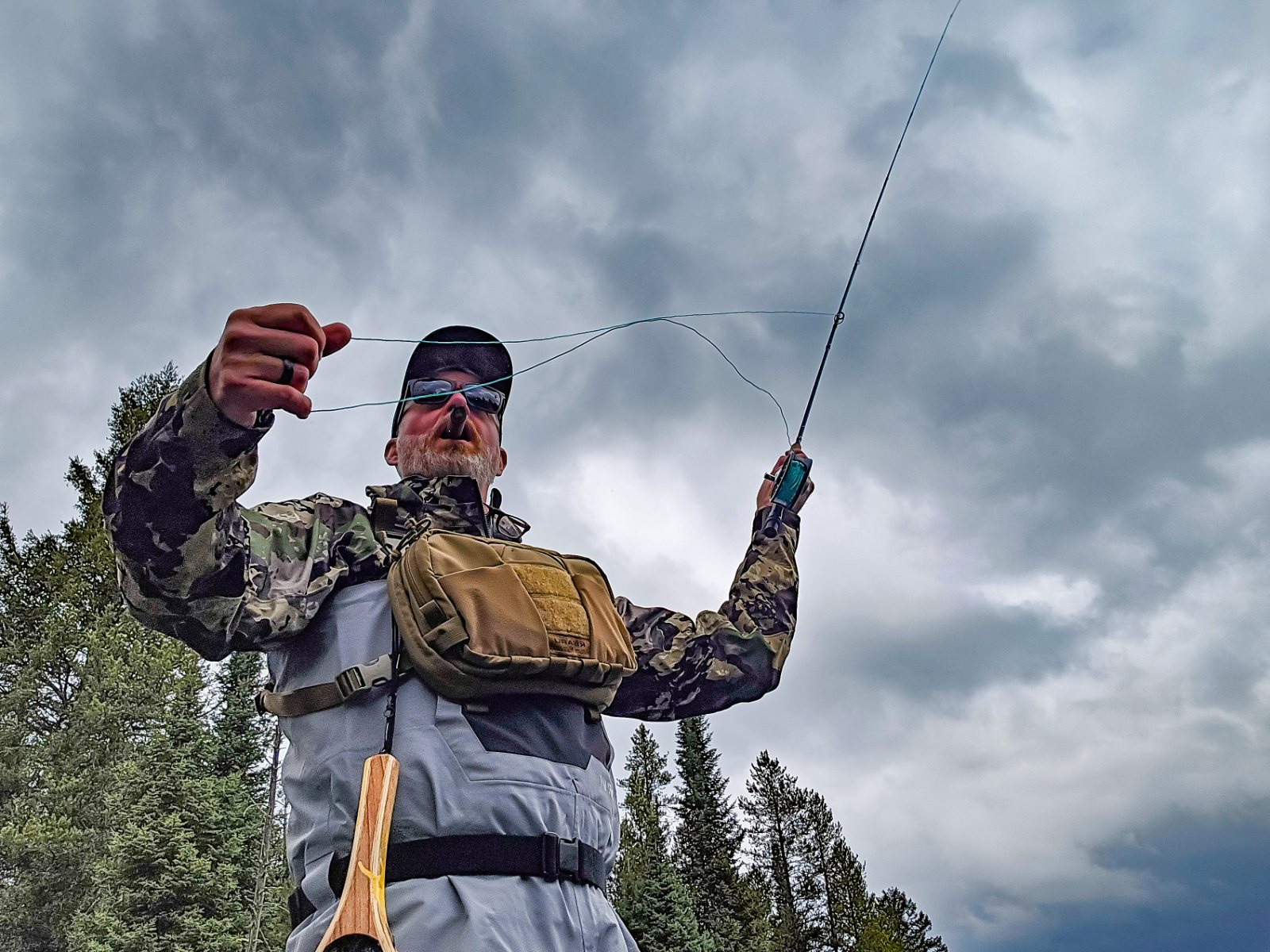 Jon Wayne Taylor casting a fly rod in waders and a Kifaru chest harness under a stormy sky